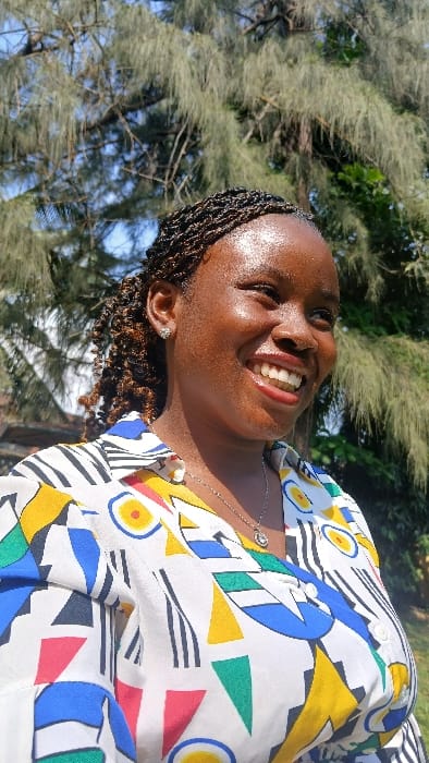 A close-up outdoor portrait taken in Awjaw, Nigeria, features an adult female with dark skin, smiling broadly and looking towards the right side of the frame. Her face shows prominent highlights, indicative of direct sunlight. She has dark, braided hair styled in small twists, some visible as curly strands. She wears small, silver-colored stud earrings in both earlobes and a silver-colored necklace with a circular pendant. Her attire consists of a white button-up shirt patterned with abstract geometric shapes in yellow, blue, red, green, and black. The background comprises green foliage from a large tree with needle-like leaves, under a clear light blue sky.