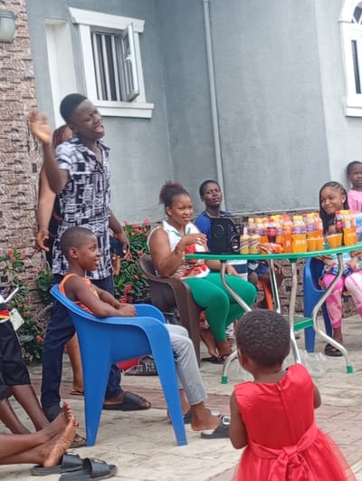 This image captures a lively outdoor gathering of people, ranging from young children to adults, in what appears to be the courtyard or patio area of a residential building in Onicha, Nigeria. The atmosphere is cheerful and relaxed, suggesting a social event or casual celebration.

In the center of the frame, a young man wearing a black and white patterned shirt stands with an expressive, open-mouthed gesture, his left hand raised, suggesting he is speaking, singing, or telling a story. Many attendees are looking towards him, engaged by his presence. To his left, a young boy in an orange tank top sits in a blue plastic chair, smiling brightly.

Further back, a green rectangular table serves as a focal point, laden with numerous bottles of juice or soft drinks in various colors, along with several cups and a few cans, one of which clearly displays "FANTA." A black speaker or boombox is also visible on the table. Around the table, several individuals are seated: a woman in a white top and green leggings smiles from a brown chair, while another young girl in a pink top and patterned leggings smiles from a blue plastic chair. A woman in a blue top is partially visible behind the table.

In the foreground, with her back to the camera, a very young child in a bright red dress adds to the multi-generational presence. Other individuals are partially visible at the edges of the frame, some sitting on the ground or in chairs, with sandals and flip-flops nearby.

The setting includes a building with textured grey brickwork on the left and a plain grey wall on the right, featuring a white-framed window with security bars, partially ajar. Green flowering bushes provide a natural element behind the seated guests. The ground is paved with light-colored interlocking stones or tiles. The bright daylight indicates it is daytime, and the overall mood conveys enjoyment and camaraderie.
