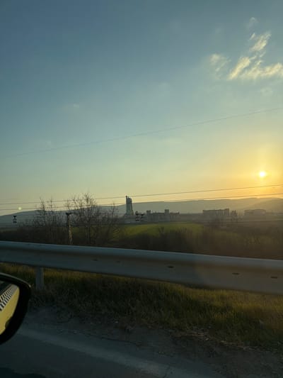 This image depicts a wide shot of an industrial and rural landscape at sunset, viewed from a vehicle on a road near Kipra, Bulgaria.
In the foreground, a metallic, silver-colored guardrail runs horizontally across the lower third of the frame, separating the viewer from the scene beyond. A paved road surface is visible below the guardrail on the left, and a portion of a car's side mirror, reflecting a patterned interior, is visible in the lower left corner. The immediate roadside verge consists of dry earth and sparse green grass.
The midground features expansive green fields with scattered, bare deciduous trees, suggesting late winter or early spring. A cable car system with multiple white gondolas on wires and support pylons traverses these fields horizontally from left to right. Overhead power lines also stretch across the frame above the fields and trees.
In the background, a large industrial complex, possibly a factory or processing plant, is visible, characterized by multiple grey/green buildings and a prominent tall, light-colored tower structure on its left side. Beyond the industrial complex, a range of hills or mountains forms the horizon, appearing hazy due to distance and atmospheric conditions.
The sky, dominating the upper portion of the image, transitions from a pale blue at the top to a bright yellow-orange gradient towards the lower right. The sun, a brilliant yellow orb, is visible in the upper right quadrant, indicating sunset or late afternoon, casting a warm glow over the entire scene. Sparse, wispy clouds are present in the blue expanse of the sky. No individuals are visible.