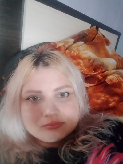 The image is a close-up, indoor selfie of a young woman with blonde hair. She is lying down, with her head resting on a pillow that has an orange and brown patterned covering, possibly a blanket or comforter. The background consists of a wall and a dark headboard. The woman is wearing makeup, including eyeliner and lipstick. Her hair is styled loosely, with some strands falling around her face. The lighting suggests an indoor setting, possibly a bedroom. The location of Изыхские Копи, Russia is not determinable from the visual information provided in the image.