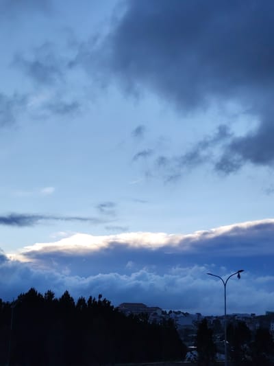 The image depicts a cityscape in Lang Biang - Da Lat, Vietnam, at dusk or dawn. The sky is a gradient of blues and grays, with a layer of clouds illuminated from behind, suggesting the sun is near the horizon. In the foreground, a dark silhouette of trees lines the horizon. A street light stands to the right, with a bent arm and a damaged lamp. Beyond the trees, a cluster of buildings, likely residential, is visible. The overall scene suggests a quiet moment at the edge of a city.