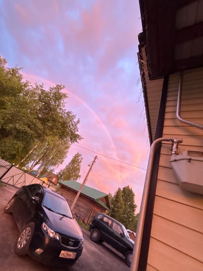 The image depicts a rural scene with a rainbow arcing across a pink and purple sky. In the foreground, a black Kia car is parked on a dirt driveway. To the right, a house with beige siding and dark brown trim is visible, with a gas meter attached to the exterior wall. Behind the car and house, trees and other residential buildings are present. The overall lighting suggests it is either sunrise or sunset. The location is identified as Zaprudnya, Russia.
