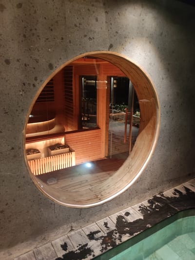 This image captures a nighttime view through an oval-shaped opening in a dark gray, speckled concrete wall, revealing a warm-toned wooden interior space and an exterior area beyond it, located in Mas, Indonesia.
**Scene Elements:**
- **Foreground (bottom right):** A light greenish-blue swimming pool is partially visible, bordered by a dark-stained wooden deck showing wet patches.
- **Concrete Wall:** The dark gray concrete wall, speckled with lighter aggregate, dominates the foreground and frames the central oval opening.
- **Oval Opening:** The opening is framed by a smooth, light-colored wooden archway.
- **Wooden Interior (visible through opening):** Inside, the walls and floor are constructed from warm-toned wooden panels and floorboards.
- **Left Side:** A circular wooden tub or basin with an illuminated base is situated on an elevated wooden platform.
- **Center:** Below the tub, two square-shaped containers filled with rocks, illuminated from within, are visible behind a vertical wooden slat barrier.
- **Back:** A clear glass door with two vertical bronze-colored handles leads from this wooden room to an exterior space.
- **Exterior Area (beyond glass door):** A dimly lit outdoor patio or garden area features dark paving and planters containing green foliage.
- **Lighting:** The interior wooden space is brightly illuminated by warm, artificial lights, casting a glow. The concrete wall is subtly lit, with a brighter patch in the upper right.
**Absence of:**
- No individuals are present in the scene.
- No direct actions or interactions are depicted.