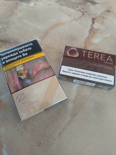 Two cigarette packs are on a patterned countertop. The pack on the left is a Davidoff Gold pack featuring a graphic health warning of a diseased mouth. Text on the pack is in Bulgarian and includes "Тютюнопушенето уврежда зъбите и венците Ви" which translates to "Smoking damages your teeth and gums". The pack on the right is a TEREA BRONZE pack for IQOS ILUMA. Text on this pack is also in Bulgarian and reads "Това тютюнево изделие уврежда Вашето здраве и води до пристрастяване", translating to "This tobacco product harms your health and causes addiction." The scene does not provide any information about the city of Yagoda, Bulgaria.
