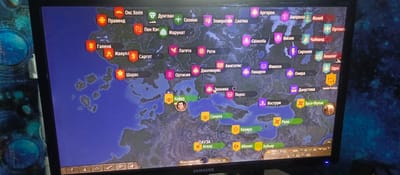 This is a photograph of a computer screen displaying a map from a video game. The map shows various regions with city names and icons. The game appears to be a strategy or simulation game, possibly set in a fantasy or historical world, given the names of the locations such as "Аргорон," "Марунат," and "Аккалат." The screen is showing a colorful map with various political territories marked by different colored banners. Some locations have crests or symbols, and others have portraits of characters, such as the one labeled "Кайяз." The bottom of the screen shows game interface elements including date and time ("Весна, 8 день, 1087 год"), resources (numbers like "1.5," "100%," "94," "253," "53.7," "10," "15.1," "1076"), and playback controls, indicating the game is currently being played or paused. The overall scene is of someone playing a video game, with the computer monitor as the central subject. The background of the screen has a dark, starry pattern, suggesting it's a game map or interface element.