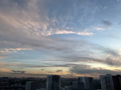 This is a wide-angle, eye-level photograph of a cityscape at dusk, with a dramatic sky filled with clouds. The foreground is dominated by several modern, multi-story apartment buildings with white and gray facades. These buildings are clustered together, forming a dense urban landscape that extends towards the horizon. In the background, more buildings of varying heights are visible, some appearing to be under construction with cranes dotting the skyline.
The sky is the most striking element of the image. It is a canvas of blended colors, transitioning from a pale blue at the top to soft oranges, pinks, and grays as it nears the horizon. Wispy, elongated clouds stretch across the sky, catching the fading sunlight and creating a textured, layered effect. Some clouds are tinged with a golden hue, suggesting the last moments of sunset, while others are darker, hinting at approaching evening or lingering weather patterns. The overall mood is serene and slightly melancholic, capturing the transition from day to night over a developing urban area. No people are visible in the image. The location context suggests this is a view of residential buildings in Бордуки, Russia.