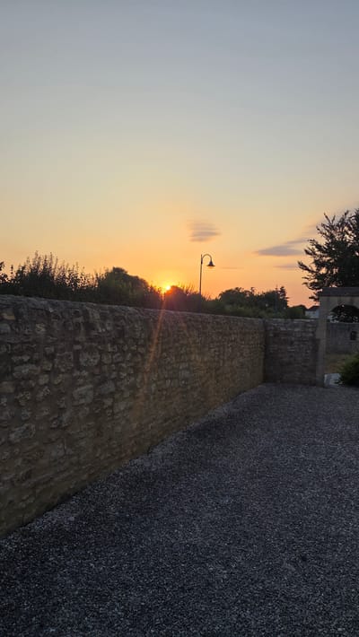 The image depicts a sunset scene in the city of Macé, France. A stone wall runs diagonally across the foreground, separating a gravel-covered area on the right from a grassy area with trees on the left. The sun is setting behind a line of trees and a street lamp is visible to the right of the sun. The sky is a gradient of orange and yellow hues, with a few wispy clouds. The overall impression is one of a peaceful, rural landscape at dusk.