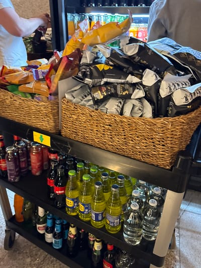 A person wearing a white t-shirt is reaching into a basket filled with various snack bags. The basket is one of two woven baskets on a black multi-tiered rolling cart. The top shelf of the cart holds the two baskets of snacks. The second shelf holds rows of bottled beverages, including what appear to be sodas and water bottles. Some of these bottles are green, yellow, red, and clear. The bottom shelf also holds bottled beverages, with some appearing darker in color, and other types of bottles. A bright yellow sign with a green arrow pointing down is attached to the front of the second shelf. The background shows what looks like a commercial establishment with shelves of drinks visible. The floor appears to be tiled. This scene is located in Callao, Peru.