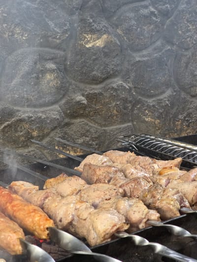 The media file shows a close-up, overhead view of meat being cooked on skewers. The meat is in various stages of cooking, with some pieces appearing more browned and others a lighter pink. The skewers are metal and are arranged vertically, with the meat threaded onto them. The meat is seasoned with black pepper. The skewers are positioned over what appears to be hot coals, as evidenced by the red glow visible beneath the bottom skewer. Wisps of smoke are rising from the meat, indicating that it is actively cooking. The background consists of a rough, textured stone wall. The lighting suggests it is daytime, and there are no indications of weather conditions. The location is identified as Pidhorodne, Ukraine. No people are visible in the frame. The overall scene depicts a barbecue or grilling event, likely outdoors given the stone wall and the cooking method. The focus is on the food and the cooking process.
