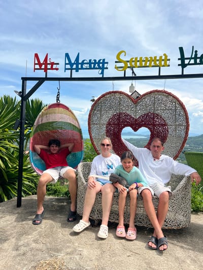 A family of four, two adults and two children, are posing for a photo in front of a large wicker heart sculpture. The family is seated on a wicker sofa. To the left of the sofa, a teenage boy is sitting in a colorful hanging egg chair. Above the family, a sign with colorful letters reads "Mr. Mong Samui Vie." The background shows a lush green landscape with mountains and a body of water, indicative of a scenic viewpoint in Baan Thurian, Thailand. The sky is partly cloudy. The family appears relaxed and happy.