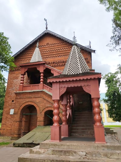 This is an exterior, low-angle, eye-level shot of a red brick building with elaborate red wooden architectural features. The building has a steeply pitched roof with decorative trim along the eaves and multiple decorative spires and finials. A prominent, ornate red wooden porch with a conical roof extends from the upper story. A staircase with red wooden balustrades leads up to this porch. Below the porch, the brick facade has arched openings and a sloping metal cover. The building is surrounded by lush green trees, and a cloudy sky is visible. The overall style of the architecture suggests a historical or cultural site, possibly a church or a museum. The image is undated and no individuals are present in the frame. The location is identified as Chelganovo, Russia.