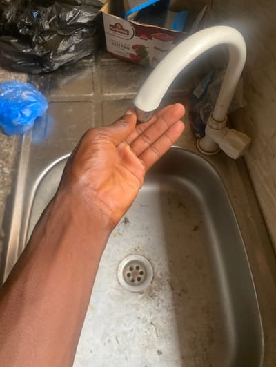 A person's arm and hand are positioned near a white faucet over a sink. The faucet is curved and appears to be made of plastic. The sink is made of stainless steel and has a drain with multiple holes. The surrounding counter appears to be made of a grey material, possibly tile or concrete, and is somewhat grimy. To the left of the sink, there is a cardboard box with red and white labeling that reads "RASHNIK CHILLI & NUT CUTTER." Below the box are black and blue plastic bags, suggesting a trash area. The lighting suggests an indoor environment, possibly a kitchen or utility area. The image is taken from a top-down perspective, focusing on the hand and faucet. There are no clear indicators of the time of day or weather. The overall impression is of a functional, perhaps slightly neglected, space.