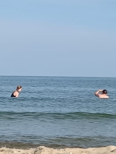 Two people are swimming in the Black Sea at Zatoka, Ukraine. On the left, a young woman with blonde hair is wearing a black swimsuit and is facing towards the right. On the right, a person with short brown hair is swimming and has their right arm raised, touching their head. The water is calm with small waves and the sky is clear and light blue. The image appears to be taken from the shore, with a sandy beach visible in the foreground.