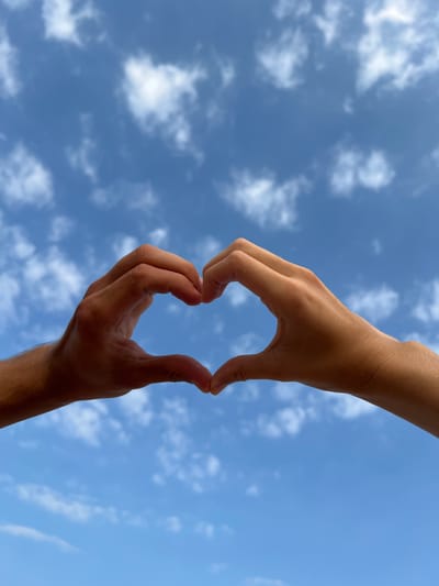 Two hands, one above the other, are positioned against a blue sky with scattered white clouds, forming a heart shape with the fingers. The lower hand is more tan, while the upper hand has a paler skin tone. The location is not identifiable as Nam Natural Heritage Area Trang, Vietnam based solely on the visual elements of the image.