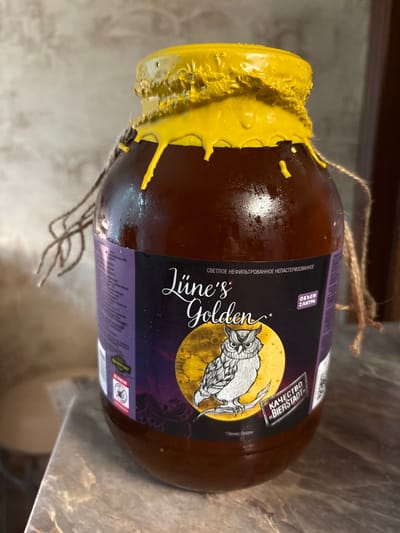 The image shows a large, brown glass bottle of "Lüne's Golden" beer, with a yellow wax seal dripping down the neck. The bottle is lying on its side on a textured surface that appears to be a countertop or table. A rustic twine is tied around the neck of the bottle, with ends hanging loose.

The label on the bottle features a dark purple background with a circular graphic depicting an owl silhouetted against a large, yellow moon. The text on the label is in Cyrillic and Latin scripts. Visible text includes "Lüne's Golden," "СВЕТЛОЕ НЕФИЛЬТРОВАННОЕ НЕПАСТЕРИЗОВАННОЕ" (Pale Unfiltered Unpasteurized), "ОБЪЕМ 2 ЛИТРА" (Volume 2 Liters), and "КАЧЕСТВО «BIERSTADT»" (Quality "BIERSTADT"). The text "Плюнес Голден" is also visible. There are warning symbols on the label, including one depicting a crossed-out cigarette.

The lighting suggests an indoor setting, likely a home or a bar, with diffused light. The overall impression is of a decorative or artisanal presentation of the beer. The yellow wax seal adds a handmade, rustic aesthetic. The background is slightly blurred, keeping the focus on the bottle. The location is identified as Pontonny, Russia.