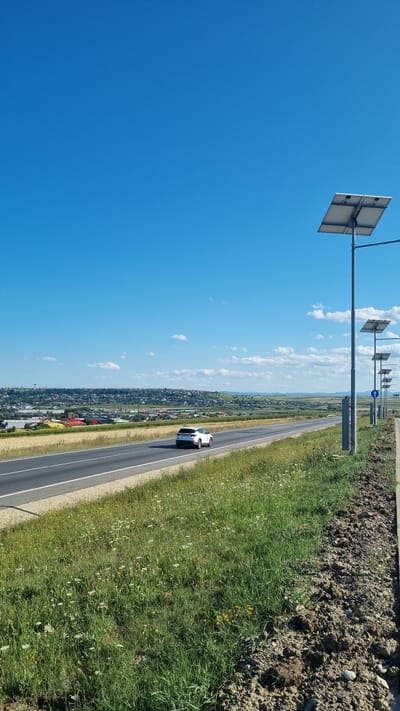 The image depicts a highway scene with a white car driving on the right lane. The highway is bordered by a grassy verge with scattered wildflowers. To the right of the highway, a series of lampposts equipped with solar panels are erected. In the background, a town with houses and some industrial buildings is visible, situated on a gentle slope under a clear blue sky with scattered white clouds. The location is identified as Șcheia, Romania.