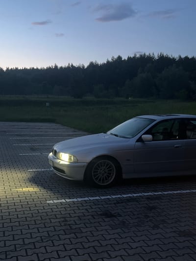 The image depicts a silver sedan parked in an outdoor paved lot. The car's headlights are illuminated, casting a bright beam onto the interlocking brick ground. The lot appears to be a parking area, indicated by white painted lines marking parking spaces. In the background, a dense line of trees forms a dark horizon against a twilight sky, transitioning from a pale blue near the horizon to a darker shade above. The overall scene suggests an evening setting in the city of Koziegłowy, Poland.