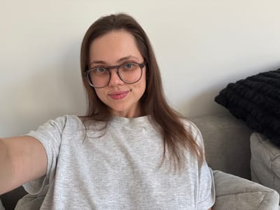 The image shows a young woman with long, brown hair, wearing glasses and a grey t-shirt. She is seated on a grey sofa in what appears to be an indoor setting, with a white wall behind her. A black textured pillow is visible on the sofa to her right. The lighting is natural, coming from the left, casting a soft light on her face and the surrounding environment. There is no indication of the city of Szklarska Poręba, Poland, in the image.