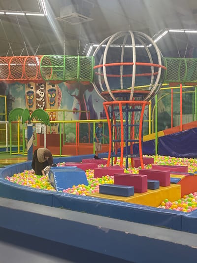 This image depicts the interior of a children's indoor play area, likely a playground or entertainment center, in Tri Protoka, Russia. The scene is vibrant and colorful, filled with various structures designed for play and physical activity.

Dominating the upper left of the frame is a large, spherical netted structure, possibly a climbing dome or observation pod, suspended from the ceiling. Below this, orange and blue netting forms part of a climbing frame or obstacle course. To the right, a ball pit overflows with numerous brightly colored plastic balls in shades of pink, yellow, and green. Several large, padded blocks in blue, pink, and yellow are arranged in a tiered fashion next to the ball pit, serving as steps or platforms.

In the background, a mural adorns the wall, featuring cartoonish depictions of creatures and stylized trees, contributing to the playful atmosphere. Green netting structures and orange support poles are visible throughout the play area, indicating multiple levels and interconnected play zones.

Two individuals are present in the scene. One person, wearing a blue t-shirt, is visible standing within one of the netted play structures. Another individual, seen from the back, is seated or kneeling near the ball pit, appearing to be engaged with the balls or resting. The overall lighting suggests it is daytime, likely indoors given the presence of artificial lighting fixtures visible in the ceiling. The environment appears to be designed for active children's recreation, with a focus on fun and safety. No visible text is present in the image.