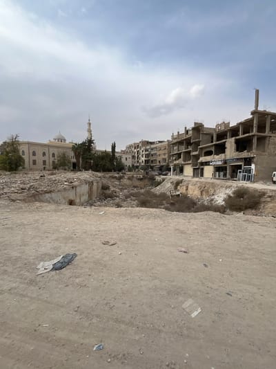 This image captures an urban landscape in Harasta, Syria, under an overcast sky. The foreground consists of an unpaved, light-brown dirt ground, scattered with debris, including a folded black and grey fabric item in the lower-left.

A large, irregular depression or excavated area dominates the middle ground, characterized by exposed earth, rubble, and scattered dry vegetation. A partially obscured white object, possibly refuse, is visible on the right slope of this depression.

In the background, to the left, stands a large, light-colored mosque featuring a central dome and a single slender minaret, surrounded by trees and appearing structurally intact. To the right, a street runs parallel to the excavated area, lined by multi-story concrete buildings. Many of these structures exhibit significant damage, with visible exposed rebar, missing walls, and incomplete floors, suggesting extensive destruction or ongoing reconstruction. Some ground-level units function as commercial storefronts; one shop on the far right displays multiple white window frames leaning against its exterior. A white panel van is parked on the street on the far right. A single individual in a light-colored garment is discernible near the window frames, and another faint figure is near the van.