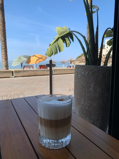 A tall glass of layered latte sits on a wooden table in the foreground. The drink is topped with a foamy white layer. In the background, a beach scene is visible through a window or barrier. Palm trees flank the view, and umbrellas are set up on the sand. People are scattered on the beach, and the ocean stretches out to the horizon under a clear blue sky. A large potted plant with broad green leaves is positioned to the right of the frame. The setting suggests a cafe or restaurant overlooking the beach in Peguera, Spain.