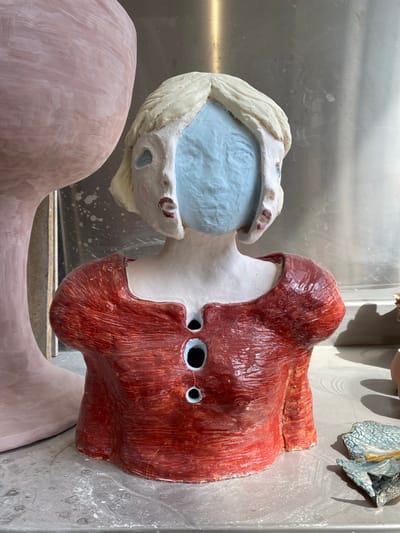 The image shows a partial, apparently unfinished ceramic sculpture. The sculpture consists of a red, roughly head-shaped form with holes for eyes, attached to a white neck leading to an open mouth with light blue inside. The mouth and the neck are a contrasting, lighter color than the red head. The sculpture is resting on another, larger, pink ceramic form, which appears to be part of the same sculpture but is less detailed. The background is a stainless-steel surface with water stains. Other ceramic pieces can be seen partially in the background. There is no evidence to suggest a specific location within Saint-Ouen-sur-Seine or any other city.