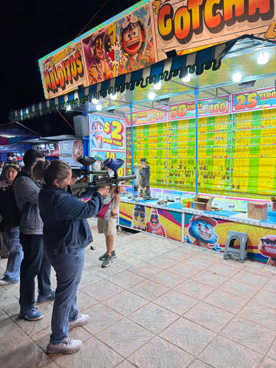 People stand at a carnival game stall, aiming paintball guns. The stall advertises "2 por tiro" and various prices for more shots, like "50 tiros por $100" and "25 tiros". The backdrop is filled with colorful targets. A sign mentions "Gotchaco". A pizzeria named "Luz" is nearby. The ground is tiled.