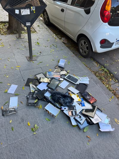 A street scene in Bahía Blanca, Argentina shows a sidewalk littered with discarded electronic devices, primarily tablets and phones, along with a black garbage bag, packaging materials, and fallen leaves. A white compact car is parked along the curb, and a black metal trash receptacle stands nearby. The sidewalk is paved with gray stone tiles.