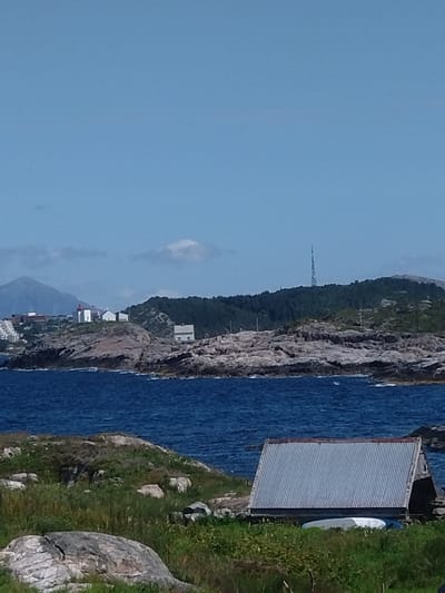 The image depicts a coastal landscape in Averøy, Norway, on a clear day with a bright blue sky. In the foreground, a grassy embankment with scattered rocks leads down to the choppy blue sea. A corrugated metal shed with a red-trimmed roof and a white boat underneath is situated on the embankment.
In the mid-ground, the sea meets a rocky coastline dotted with green vegetation. A small cluster of white buildings, including one with a red roof, sits atop the rocks. Further inland, a dense, dark green forest covers rolling hills. A tall, slender radio tower is visible on the highest point of the forested hill. In the background, hazy mountains rise against the sky, with some lighter, cloud-like formations. A hint of a town or development with white buildings can be seen on the lower slopes of the leftmost mountain.