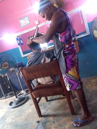 The image shows a barber giving a haircut to a customer in what appears to be a small barbershop. The barber, a young man, is wearing a colorful patterned top and shorts, a grey apron, and flip-flops. He is standing and focused on cutting the customer's hair with clippers. The customer is seated in a wooden chair and covered with a grey cape. A mirror hangs on the wall behind them, framed in white. To the left, a standing fan is visible. The wall is painted with a two-tone color scheme of pink and blue. A price list for the Orlu Barbers Welfare Union is displayed on the wall, implying the location is within the city of Orlu, Nigeria. The floor appears to be concrete.