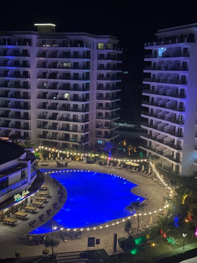 The image captures a nighttime aerial view of a resort or hotel complex, likely in Gastria, Northern Cyprus. Two large, multi-story white buildings with numerous balconies dominate the scene, illuminated by ambient light and some colored lighting. Between the buildings and extending towards the foreground is a large, irregularly shaped swimming pool with intense blue lighting.

Surrounding the pool is a paved area with numerous lounge chairs and umbrellas, some of which are occupied by unseen individuals. String lights, emitting warm yellow light, are draped around the pool area, creating a festive atmosphere. To the left, a building with a curved facade features outdoor seating with tables and chairs, and a sign that reads "Mia Bella". This building is also illuminated, with some windows showing interior lights.

The landscape surrounding the pool area includes palm trees and other vegetation, with decorative colored lights visible in some areas. The overall impression is of a lively and well-lit resort at night, with clear skies overhead. The buildings exhibit modern architecture with repetitive balcony designs. The presence of lounge chairs and umbrellas suggests a place for relaxation and recreation.