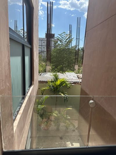 View from a window in Mérida, Mexico, looking out onto a small courtyard. The architecture is modern with bare concrete walls. A small palm tree and other foliage are visible in the courtyard. There are unfinished construction elements, including rebar, visible in the background. The sky is partly cloudy.