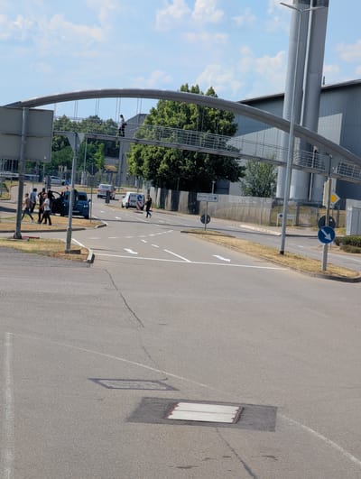 The image depicts an outdoor scene on a sunny day, likely in Sindelfingen, Germany, given the text visible on a street sign. The primary focus is a paved road with white lane markings and arrows indicating traffic flow. A prominent feature is a large, curved metal structure that appears to be an overhead walkway or part of a bridge, supported by substantial metal pillars.
Several people are visible in the scene. Some are walking on the road near a crosswalk, while others are on the elevated walkway, observing the surroundings or moving across. There are also vehicles present, including a dark blue car and a white van with visible text that seems to be "RÜM" and "RIESCHLING".
Traffic signs are also noticeable. One sign shows a blue circle with a white arrow pointing to the right, indicating a mandatory direction. Another sign has a yellow diamond shape, often used for warning signs. A street sign reads "Mercedes-Benz Tor 40", suggesting proximity to a Mercedes-Benz facility, which aligns with the location context.
The weather appears clear and sunny, casting shadows on the road and the walkway. The time of day is likely daytime, given the brightness and the shadows. The overall atmosphere seems to be that of a functional urban or industrial area, with elements of infrastructure, transportation, and pedestrian movement.