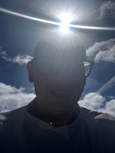 A selfie taken outdoors with the sun directly overhead, creating a strong glare and obscuring the face of the subject. The subject, seen from the shoulders up, is wearing glasses and a dark-colored shirt. The background consists of a bright blue sky with scattered white clouds, suggesting it is a sunny day. The perspective is low-angle, looking up at the subject and the sun. There are no discernible landmarks or context to place this image within the city of Tewkesbury, United Kingdom.