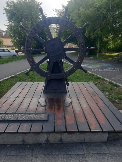 The image shows a monument in the city of Shelekhov, Russia. The monument is a large, dark metal ship's wheel mounted on a wooden platform. The wheel has handles and is decorated with a crest in the center. Inscriptions are visible on the wheel and on a plaque attached to the wooden platform. The wooden platform is made of planks with visible nails and has some red paint or staining. Two small, flat objects, possibly metal cutouts of footprints, are placed on the platform. The monument is situated outdoors, with a paved walkway, grassy areas, trees, and buildings visible in the background. The sky is overcast. The plaque on the platform reads "ЗДРАВСТВУЙ ГОРОДОМ В КОТОРОМ ТЫ ЖИВЕШЬ" which translates to "WELCOME TO THE CITY WHERE YOU LIVE." The ship's wheel itself has text inscribed around its rim, partially visible, including "ШЕЛЕХОВ" (Shelekhov) and "1962" indicating the city's founding year. The overall scene depicts an outdoor public space with a prominent monument.