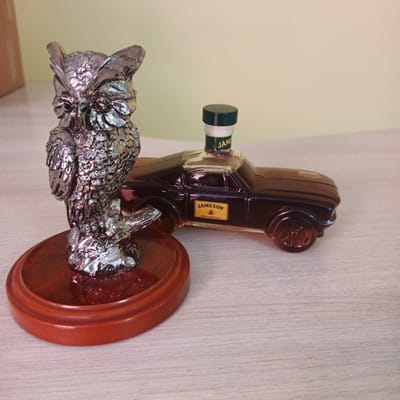 A silver owl figurine on a polished wooden base sits to the left of a dark brown, car-shaped bottle of Jameson Irish Whiskey. The bottle has a green cap and a yellow label with the Jameson logo. The items are placed on a light-colored wooden surface, possibly a table or shelf. The background is a plain, light yellow wall. The image appears to be an indoor shot, with soft lighting. There are no people or animals present. The image does not provide any information about the location in the city of Britovo, Russia.