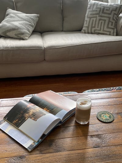 This image depicts an indoor scene within a residence in Ridgefield, United States. The upper portion of the frame is dominated by a light-beige or light-grey fabric sofa, featuring two visible seat cushions and three back cushions. Two throw pillows are present on the sofa: one on the left with a grey chevron or zigzag pattern, and one on the right with a grey labyrinth or maze pattern.
In the lower half of the frame, a dark-stained wooden coffee table with a visible grain is centrally positioned. On the table, two open books or magazines are stacked. The top publication is open to a page displaying an architectural image on the left page and text on a light background on the right page. The bottom publication is partially visible underneath. To the right of the publications, a clear glass contains a light brown, layered beverage with a frothy white top, indicative of a coffee or milky drink. A circular coaster, green with an intricate gold design, is located to the right of the glass.
The floor beneath the coffee table is composed of dark-stained hardwood planks. A multi-colored patterned rug is partially visible beneath the coffee table and books. Natural light appears to illuminate the scene, casting soft shadows. No individuals are present in the image.