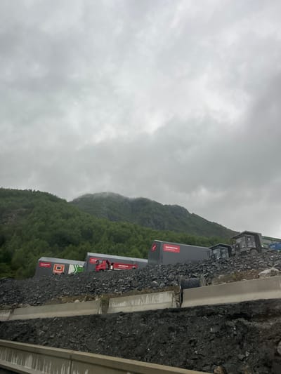 The image depicts an outdoor scene in Røldal, Norway, on a cloudy day, likely in the late afternoon. The foreground is dominated by a rough, rocky terrain interspersed with some vegetation. A concrete structure runs along the right side of the frame, suggesting a constructed area or roadway.

Further back, several large, grey modular buildings or containers are lined up. These structures have prominent red and white branding. One building clearly displays the text "METROSTU" in red, followed by smaller text below. Another building features similar "METROSTU" branding. Visible on one of the containers is the text "NAHOGSN", also in white on a red background. There's also a red truck with a white cab visible between two of the grey containers, with partial text "METRO" on its side.

Behind the buildings, a steep, densely forested mountain slope rises, shrouded in mist and cloud cover, indicating damp weather conditions. The overall atmosphere is overcast and somewhat subdued, consistent with a cloudy day in a mountainous region. No people are visible in the image. The scene suggests a construction or industrial site situated in a natural, rugged environment.
