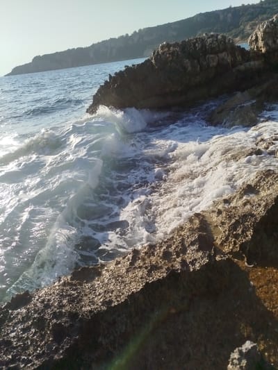 A close-up, low-angle shot captures a crashing wave against rugged, sun-drenched rocks on the coast of Karaburun, Turkey. The water is frothy and white as it breaks, with sunlight glinting off the surface, creating a dynamic and energetic scene. The rocks, in the foreground and mid-ground, are textured and appear to be porous, with varying shades of brown and gray. In the background, the calm, blue sea stretches towards a distant, tree-covered hillside under a clear, pale sky. No people or animals are visible in the frame.