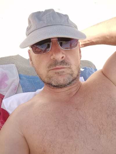 A man is lying on a beach in Kranevo, Bulgaria, holding his arm behind his head. He is wearing a grey baseball cap and sunglasses. The man is shirtless and his chest is covered in dark hair. He is lying on a beach towel with a blue and white stripe pattern. Behind him, there are other beachgoers and beach umbrellas visible. The sand is light brown and appears to be wet, suggesting the man is close to the water. The lighting suggests it is a sunny day.