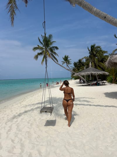 A tropical beach scene features a woman in a bikini standing on white sand near a swing hanging from a palm tree. Clear turquoise water meets the shore, where other people are visible. Palm trees line the background, some providing shade with thatched-roof structures. The sky is clear and blue, suggesting a sunny day.