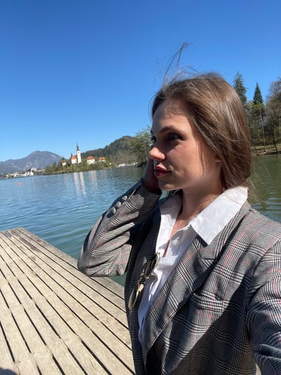 A young woman is seated on a wooden dock beside Lake Bled, Slovenia. She is wearing a white collared shirt under a grey and black plaid blazer. The lake is behind her, reflecting the clear blue sky and a mountainous landscape. In the distance is Bled Island with the iconic Pilgrimage Church of the Assumption. The woman is looking down, and her hair is slightly windswept. Sunglasses are hanging from the blazer.