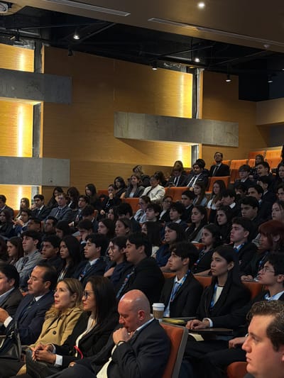 The image displays an indoor auditorium or conference hall located in Fraccionamiento Los Heroes Queretaro, Mexico. Approximately 70 to 80 individuals are seated in tiered rows of orange-brown upholstered chairs, facing forward towards an unseen focal point. The audience primarily consists of young adults and students, with a visible minority of older adults. Most attendees are dressed in business formal attire, characterized by dark suits, blazers, white or light-colored shirts, and ties. Multiple individuals are wearing lanyards with identification badges around their necks.
The room's architecture includes light wood-paneled walls structured with intersecting vertical and horizontal sections, some of which are internally illuminated, creating glowing light strips. Darker, rectangular panels, possibly concrete or metal, are integrated into the wall design. The ceiling is dark and exposed, featuring an array of track lights and linear recessed lighting fixtures. Each seat appears to have an attached small writing surface. A disposable coffee cup and a mobile phone are visible on the tables in the foreground. The overall scene suggests a formal event, such as a conference or lecture.