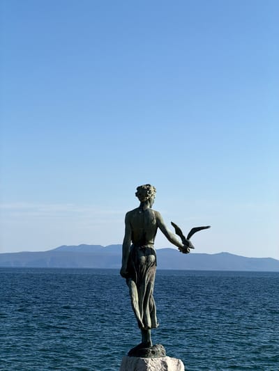 The image depicts a bronze statue of a man holding a seagull in his hands. The statue appears to be located on a stone base at the edge of the sea. The background includes the blue sea and a distant coastline. This is a view of the Maiden with the Seagull statue at Grad Opatija, Croatia.