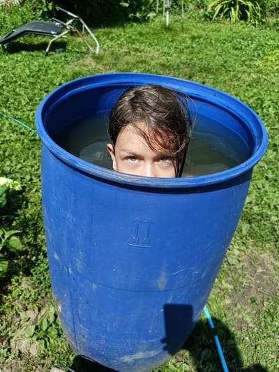 In Lagovskoe, Russia, a young person is submerged in a large blue plastic barrel filled with water. They are looking directly at the camera with their eyes and forehead visible above the water's surface. The barrel is situated outdoors on a grassy area, with greenery and some discarded items visible in the background, suggesting a casual, possibly domestic, setting. The lighting indicates it is daytime, likely sunny.