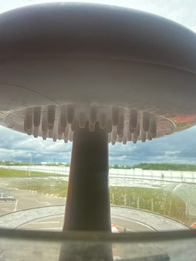 This is a close-up, slightly angled interior shot of what appears to be a shower head. The shower head is a light-colored, mushroom-shaped dome with a series of white, cylindrical protrusions on its underside. A darker, cylindrical stem extends downwards from the center of the dome, suggesting it's attached to a shower arm.

Through the transparent shower head and the background, a cloudy sky is visible, with a distant landscape of flat terrain, possibly an airport or industrial area, characterized by a white fence and a strip of green. The area appears to be outdoors and potentially damp, with some reflective patches on the ground. The image provides no specific landmarks or identifiable features to pinpoint a location in Sof'ino, Russia, or any other city. There are no people or discernible actions taking place. The scene is static and focuses solely on the details of the shower head and its immediate surroundings as viewed from within.