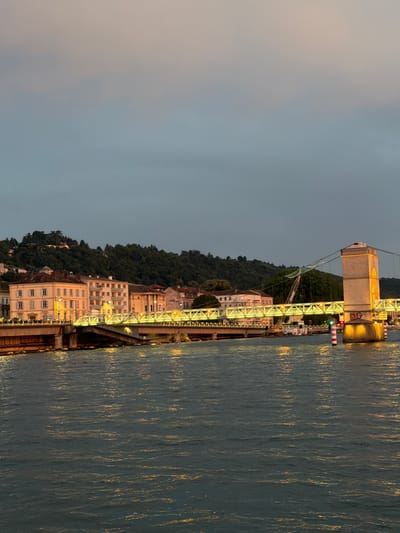 The image captures a twilight scene of a riverfront with a bridge and buildings. The perspective is from the river, looking towards the shore. Dominating the scene is a large, illuminated bridge structure, a significant portion of which is visible, including a stout, light-colored tower and a complex lattice of yellow lights that form the bridge's pathway. This bridge appears to be a pedestrian or vehicular bridge, given its scale and construction.

Along the riverbank, a line of buildings with illuminated windows suggests an urban setting. These buildings vary in height and architectural style, with some appearing older and others more modern. They are nestled against a densely wooded hillside that rises steeply behind them. The trees on the hillside are dark, indicating the low light conditions of the evening.

The river itself occupies a significant portion of the right side of the frame. Its surface is dark blue-green, reflecting the ambient light and the lights from the bridge and buildings, creating shimmering golden streaks. The water appears calm, with gentle ripples.

The sky above is a gradient of muted grays and pale oranges, characteristic of dusk or dawn. The lighting from the bridge and buildings casts a warm, golden glow on the scene, creating a peaceful and somewhat romantic atmosphere.

No people are clearly visible in the image. The activity implied is the general ambiance of a city at dusk, with the bridge serving as a prominent landmark and the buildings indicating settled habitation. The overall impression is one of a serene evening by the water in a European city, likely Vienne, France, given the location context. There is some graffiti visible on the side of the bridge tower.