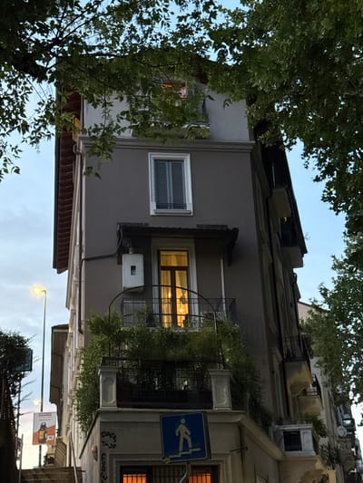 A low-angle, eye-level shot captures a narrow, tall building in Sesto San Giovanni, Italy, partially obscured by lush green tree branches overhead. The building, painted a muted gray, features a prominent dark-sloped roofline on the left side, with visible reddish-brown eaves. Several windows dot the facade; one on the upper floor is a tall, rectangular unit with a white frame and dark shutters. Below this, a balcony with black metal railings encloses a brightly lit window, revealing warm interior lighting and what appears to be a door with glass panels. An electrical box is mounted on the building's exterior wall to the left of this balcony.
To the left of the main building, a tall, slender streetlamp with a yellow light illuminates the scene. Further down, a large rectangular billboard displays an image of a yellow object, possibly a speaker or battery, on a white background.
The lower portion of the building reveals more architectural details. A railing extends horizontally, supporting planters overflowing with greenery. Below this, another balcony features a white decorative pillar. A blue pedestrian crossing sign with a white silhouette of a person walking is affixed to the wall. Further down, graffiti is visible on the building's exterior. The overall impression is of an urban environment with mature trees providing a natural canopy.