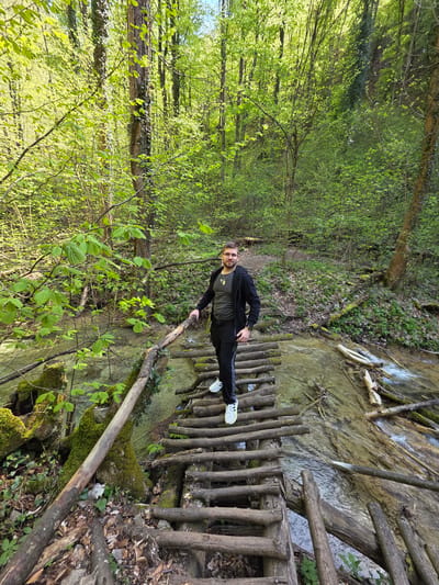 A man is standing on a wooden bridge in a forest. The bridge spans a stream. He is wearing a black jacket, a gray shirt, and black pants, and white sneakers. He is looking at the camera. The forest is green with lush trees. The water appears clear. The location is near Бачковски манастир, Bulgaria.