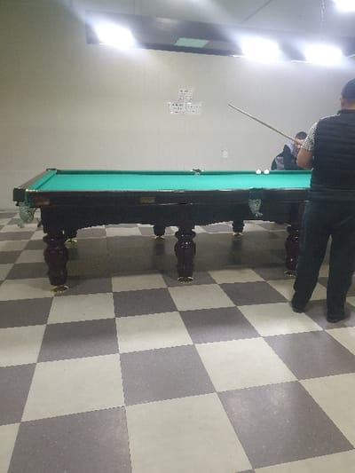 A man in dark clothing stands by a pool table, cue in hand, ready to strike the cue ball. The table, with its green felt surface and ornate legs, sits on a checkered floor. The wall is light, featuring a window and a notice board with papers. The room appears well-lit, with a stark light source shining from the top left.